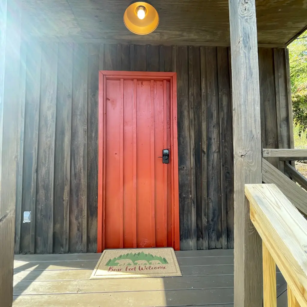 Rustic Bunk Cabin porch with red door and Bear Feet Welcome mat – Rustic Bunk Cabin at The Outpost NRG